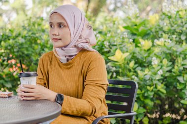 Genç Endonezyalı Müslüman bir kadın parkta oturmuş masada bir fincan buzlu kahveyle