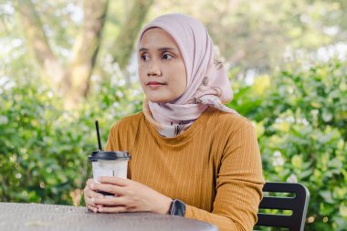 Genç Asyalı Müslüman bir kadın parkta oturmuş masada bir fincan buzlu kahveyle