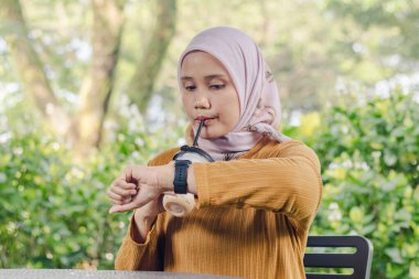 Güzel bir Asyalı Müslüman kadın buzlu kahvesinin tadını çıkarırken saatini kontrol ediyor.