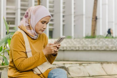 Endonezyalı genç bir müslüman kadın parkta otururken akıllı telefonuyla kayıyor.