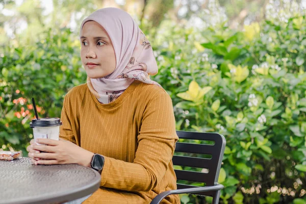 Genç Endonezyalı Müslüman bir kadın parkta oturmuş masada bir fincan buzlu kahveyle