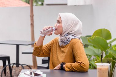 Genç Endonezyalı Müslüman bir kadın kahve masasında otururken şişe su içiyor.