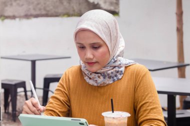 Endonezyalı Müslüman bir kadın açık bir kafede tablet üzerinde çalışıyor.