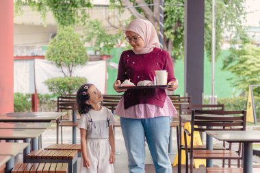 Genç bir Endonezyalı Müslüman anne ve küçük kızı birlikte yürüyorlar ve fast food restoranında tepside yemek taşıyorlar.