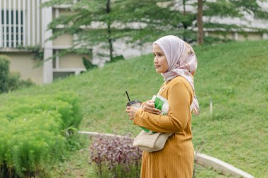 Parkta yürürken içki ve atıştırmalık taşıyan Endonezyalı genç bir Müslüman kadın.