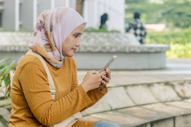 Parkta cep telefonuyla oynayan Endonezyalı genç bir Müslüman kadın.