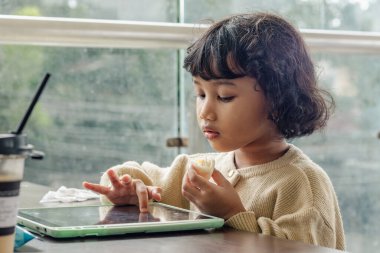 Küçük tatlı bir Endonezyalı kız tabletinde yemek yerken oyun oynuyor.