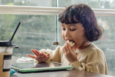 Küçük bir Endonezyalı kız tabletinde oyun oynarken yemeğin tadını çıkarıyor.