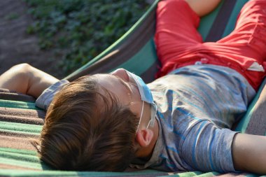 Genç bir çocuk hamakta koruyucu bir maske takarak yatıyor. Covid-19 salgını sırasında karantinaya alınmış..