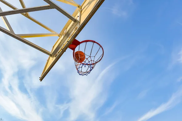 A basketball ball flies into a hoop with a net against a blue cloudy sky. Sports activities on the playground