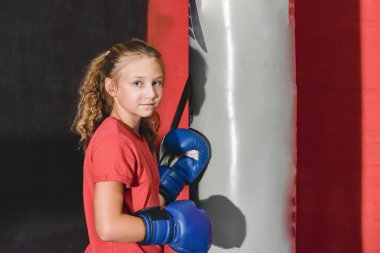 Boks eldivenli bir kız spor salonundaki bir kum torbasının arkasında duruyor..