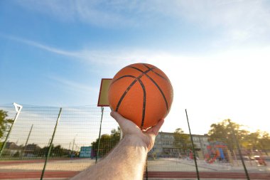 Erkek bir sporcunun eli, kalkanın arka planında yüzüklü bir basketbol topu tutuyor. Çocuk parkında spor aktiviteleri