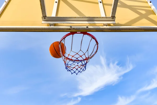 A basketball ball flies into a hoop with a net against a blue cloudy sky. Sports activities on the playground