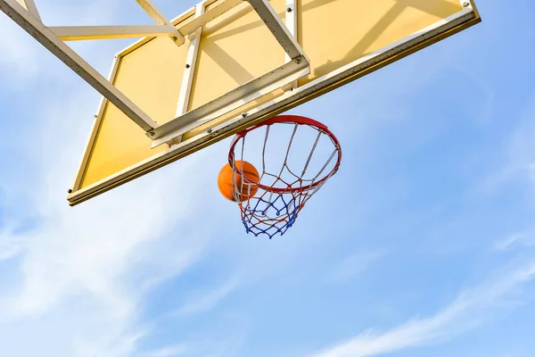 A basketball ball flies into a hoop with a net against a blue cloudy sky. Sports activities on the playground