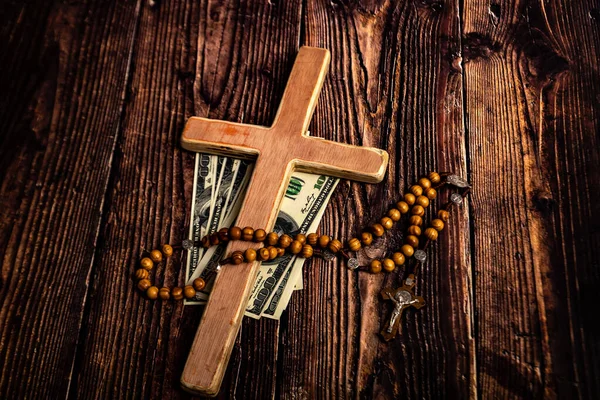 A Catholic cross, a rosary with beads and dollars lie on a dark brown wooden table.