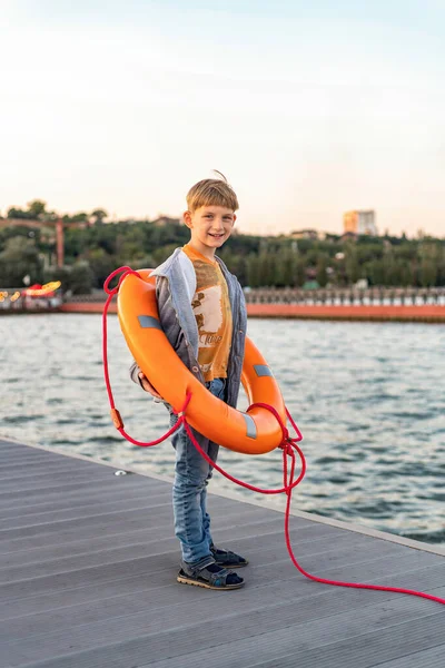 一个男孩站在海边的一个码头上 戴着橙色的救生圈 — 图库照片