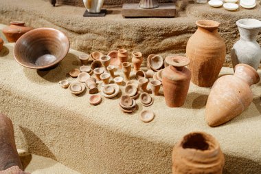 Tarihi bir yer içindeki kumların üzerinde antik çömlek parçaları sergileniyor. Objeler şekil ve boyut olarak farklılık göstererek, karmaşık işçiliği ve kültürel önemi vurguluyor..