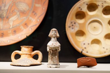 Bazı antik eserler bir müzede düzenlenir. Ortadaki süs, dekoratif çömlek ile çevrili küçük bir heykelcik. Bu kurulum tarihsel işçiliği vurgular.