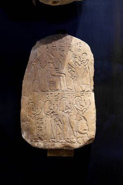Antik Mısır 'dan gelen detaylı bir taş tablet, bir firavunun ayinlerle meşgul olan hizmetkarlarla çevrili bir tahtta oturduğunu gösteriyor. Hiyeroglifler taşı süslüyor, kültürel önemi ortaya koyuyor..