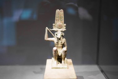 Antik Mısır 'dan gelen detaylı bir bronz heykel oturmakta olan bir tanrının asa tuttuğunu gösteriyor. Bir vitrinde duruyor, karmaşık tasarımını vurguluyor. Heykel zengin kültür tarihini yansıtıyor..