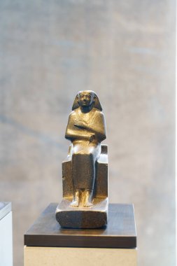 Antik Mısırlı bir figürün bronz heykeli modern bir müzede bir heykelin üzerinde dikilir. Işık, parlak yüzeyinden ince bir şekilde yansıyarak bu tarihi sanat eserinin karmaşık detaylarını vurgular..
