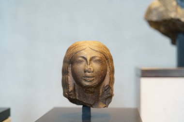 Bir kadının kafasını temsil eden taş bir heykel bir sanat galerisinde öne çıkıyor. Sergide çağdaş bir arkaplan karşısında antik sanatlar sergileniyor.