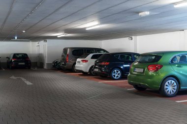 İyi ışıklandırılmış, yeraltı otoparkına çeşitli arabalar park edilmiş. Tuğla zemine çıkış yönünü gösteren bir yön oku çizilmiş..