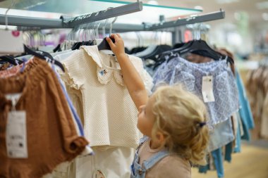 Bir mağazada, sarı saçlı, tulumlu genç bir çocuk elbise askılığında asılı beyaz bir gömleğe bakıyor. Küçük kız elbiseye uzanıyor..