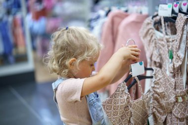 Sarışın, kıvırcık saçlı genç bir çocuk bir mağazada dikiliyor, dikkatlice giysilere bakıyor. Askıda geometrik parmak izi olan bir elbise tutuyor..