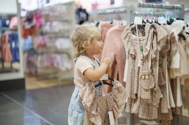 Macy 's' de, sarı kıvırcık saçlı genç bir kız askıda elbise tutuyor. Çocuk Bölümünde duruyor. Etrafı giysi parçalarıyla çevrili..