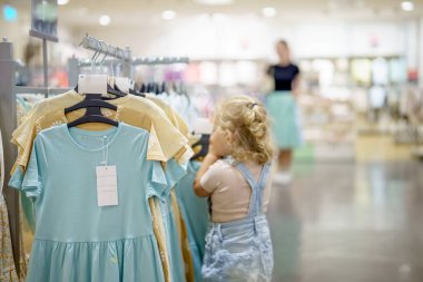 Genç sarışın bir kız, tulum giymiş, parlak ışıklandırılmış bir giyim mağazasında askıda asılı çocuk elbiselerine bakıyor. Arka planda bir kadın yürüyor, kısmen bulanık..