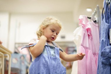 Sarışın, kıvırcık saçlı bir çocuk, bir askıdaki mavi elbiseyi inceliyor, alışveriş merkezindeki bir giyim mağazasında renkli elbiseleri inceliyor..