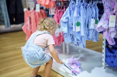 Tulumlu bir çocuk perakende mağazasındaki askıda giysileri inceliyor. Yerden bir şeyler alıyor gibi görünüyor. Giysiler sırayla asılıyor ve sahne ışıl ışıl aydınlanıyor..