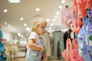 Kot pantolon giymiş sarışın bir çocuk bir mağazada dikilip elbise raflarına bakıyor. Işıklar parlak ve giysiler rengarenk. Düşünceli ve düşünceli görünüyor..