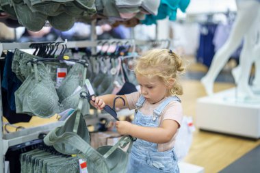 Parlak ışıklı bir mağazada, sarı saçlı genç bir kız askıdaki bir giysiyi dikkatle inceliyor. Benzer eşyalarla dolu bir rafın yanında duruyor, meraklı doğasını gösteriyor..