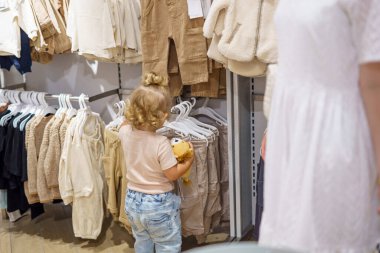 Doldurulmuş bir hayvan tutan küçük bir çocuk elbise seçimini bir giyim mağazasının rafında düşünür. Beyaz elbiseli bir kadın yakınlarda duruyor..