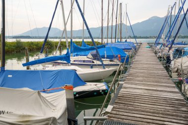 Yelkenliler yıpranmış ahşap bir rıhtım boyunca küvetlerinde dinlenirler. Tegernsee Gölü 'nün durgun suları bulutlu gökyüzünü ve Bavyera, Almanya' nın uzak dağlarını yansıtır..