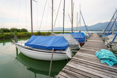 Birkaç yelkenli, sakin bir su kütlesinin üzerinde ahşap bir rıhtımda oturur. Tekneler mavi ve gri muşambalarla kaplı. Tegernsee 'de bulutlu bir günde dağlar uzaktan görülebilir..