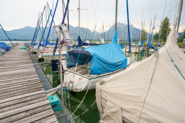 Tekneler Almanya 'da Tegernsee Gölü' nde bir marinaya yanaştı. Tekneler branda ve mavi muşambalarla kaplıdır. Dağlar bulutlu bir gökyüzünün altında arka planda yükselir ve sakin bir manzara yaratır..