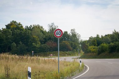 Aydınlık bir günde, bir yol bir tarlanın yanında yavaşça kıvrılır. Saatte 60 kilometre hız sınırı işareti belirgin bir şekilde duruyor, yemyeşil ağaçlar bir zemin oluşturuyor.