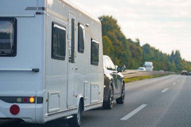 Beyaz bir karavan yolda çekiliyor, muhtemelen uzun bir yolculukta ya da tatilde. Yolculuk gündüzleri diğer araçlarla birlikte otoyolda gibi görünüyor..