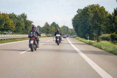 Aydınlık bir günde iki kişi motosikletlerine biner. Asfalt yol, ağaçlar ve bitki örtüsüyle çevrili olarak ileride uzanıyor. Arabalar da aynı yöne gidiyor..