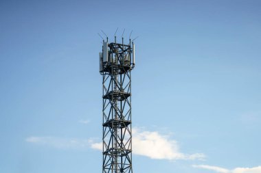 Uzun, modern bir iletişim kulesi, birkaç ince bulutla benekli açık mavi bir gökyüzüne karşı duruyor. Yapı çelikten yapılmıştır ve antenleri vardır..