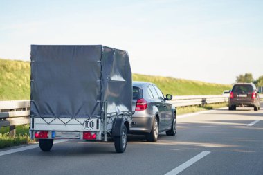 Gri renkli kompakt bir araba, kapalı bir römorku çekiyor. Uzaktan başka bir araba görünüyor. Gökyüzü parlak mavi. Güneşli bir günde yol kenarında yeşillik çizgileri.