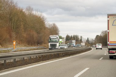 Büyük bir yük kamyonu da dahil olmak üzere bir dizi araç, çok şeritli bir Autobahn boyunca seyahat ediyor. Uzakta küçük beyaz bir minibüsün sürdüğü görülüyor. Ağaçlar otoyolun her iki tarafında da görülebilir..