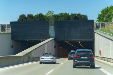 Araçlar iki yolu birbirine bağlayan parlak bir tünele doğru gidiyorlar. Tünel doğal yeşilliklerle çevrili ve gökyüzü parlak ve berrak..