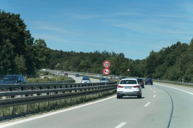 Arabalar açık mavi bir gökyüzünün altında yeşil ağaçlarla çevrili bir otoyolda sorunsuz sürüyorlar. Hız limiti işareti saatte 120 kilometre hız olduğunu gösterir..