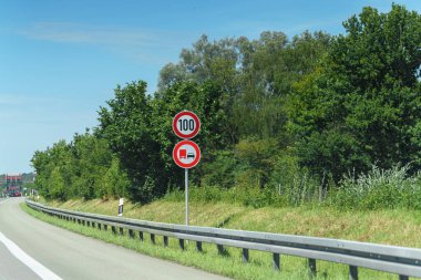 Hız limiti işareti bir kamyon sembolünün yanında, araçlar için saatte 100 km hız limitini gösterir. Yolu çevreleyen yemyeşil ağaçlar parlak bir günde canlı ve yeşil bir manzara yaratıyor..