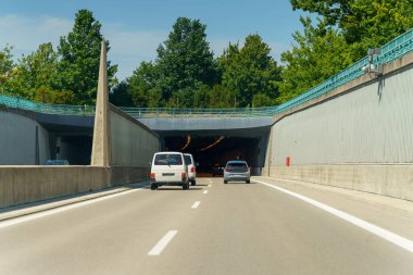 Araçlar yemyeşil ağaçlarla çevrili bir tünel girişine yaklaşıyor. Parlak mavi gökyüzü sahneye neşeli bir atmosfer ekler, tipik bir sıcak ve açık gün..