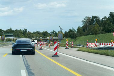 Araçlar inşaat çalışmalarının sürdüğü bir otoyolda ilerliyorlar. Yol boyunca parlak güvenlik bariyerleri var, bu da yolu uzattığımızı gösteriyor. Gökyüzünde bulutlar olan açık bir yaz günüdür..